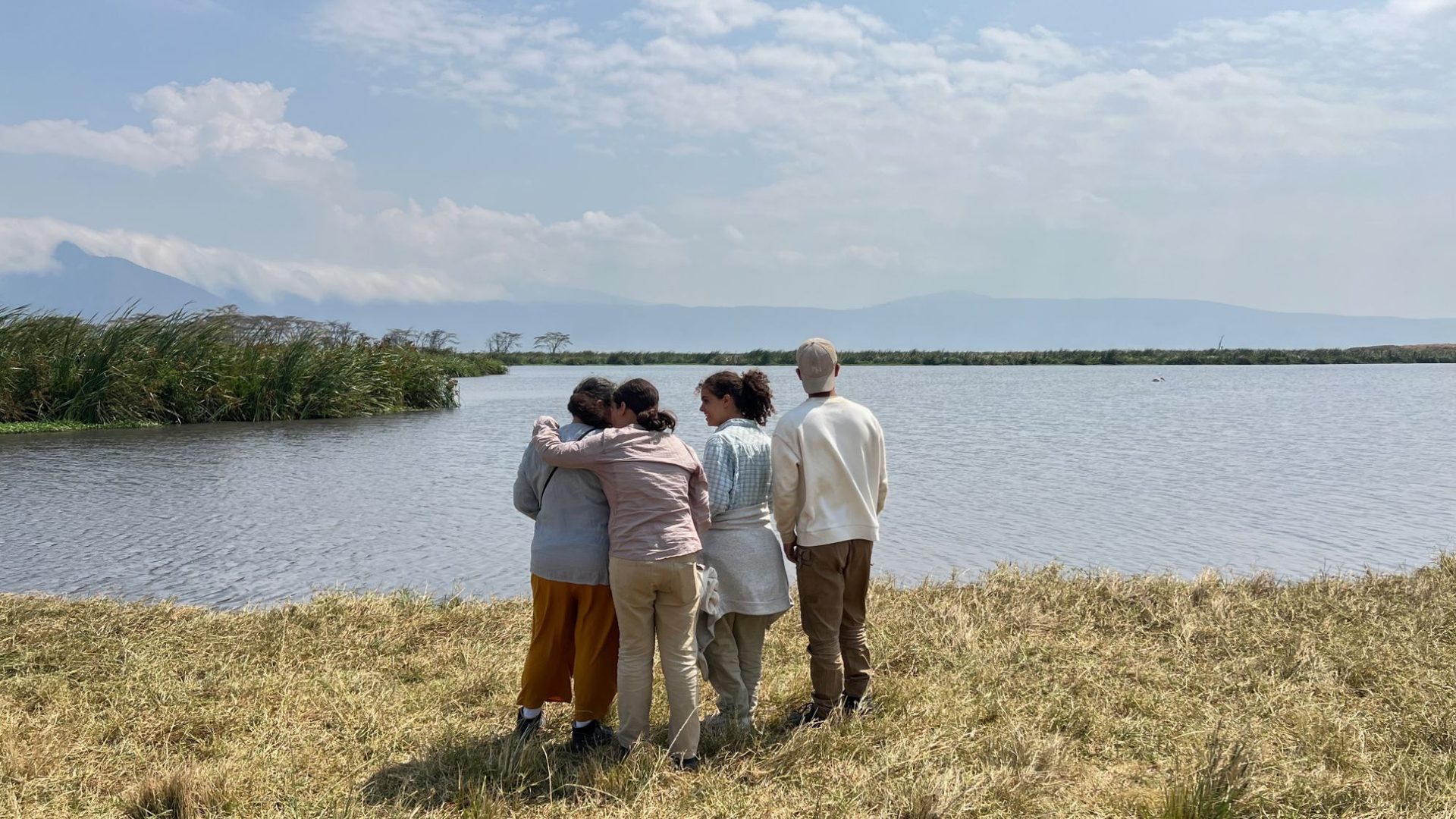 A family standing together at a crater-floor lakeshore, arms around each other, watching the water with the highlands rising hazy in the distance.